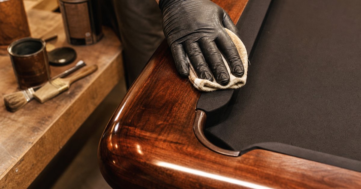 Close view of a finisher's gloved hand rubbing a soft cloth along the glossy walnut rail of a billiard table in a workshop, warm light reflecting off the deep grain, cans and brushes on a bench nearby