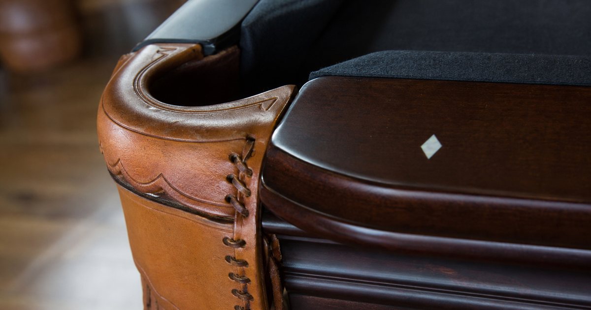 Macro photograph of a hand-laced tan leather corner pocket with fringe on a dark mahogany billiard table, a mother of pearl diamond inlaid in the rail beside it, soft window light